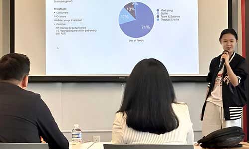 A student speaks into a microphone while presenting a project in front of a projection screen showing a pie chart