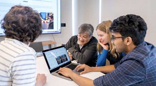 Khoury faculty member David Bau (second from left) sits at a table with three students as they view a laptop screen and discuss a research project