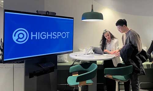 Two people discuss a project standing at a table next to a large screen with the Highspot logo