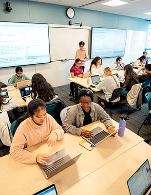 Two students sitting at a rectangular table with open laptops discuss a project while sitting in a Khoury classroom