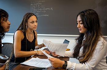 Three Khoury students sit at a table discussing a project. The student on the right is reading from a paper.
