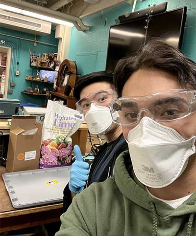 Two Griddy team members wearing ventilation masks and safety goggles in front of a table with a bag of fertilizer