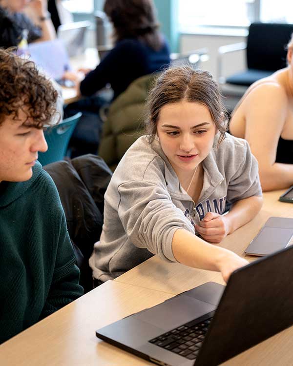 A student points at a laptop sitting on a large table as another student looks at the screen