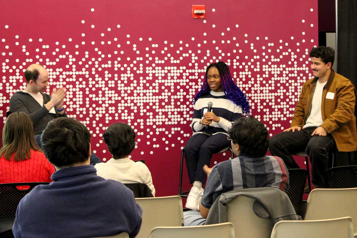Two Khoury students sit in chairs as part of a panel discussion as the audience applauds them