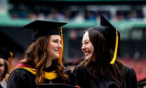 Two Northeastern graduates wearing caps and gowns smile at Fenway Park