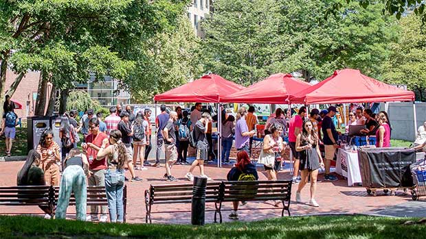 A wide view of the student clubs fair at Northeastern. Three red canopy tents are set up in a campus plaza shaded by trees. Club members stand behind tables under the tents while students interested in joining clubs ask questions.