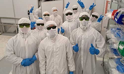 Nine Khoury College students in Silicon Valley wearing protective suits, latex gloves, and dark glasses pose for a photo during a tour about microchip production