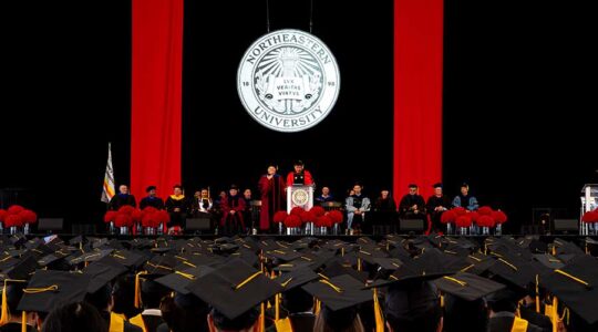 A wide view of several rows of graduates watching Ben Hescott speak from the graduation stage