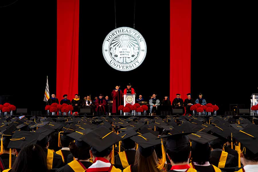 A wide view of several rows of graduates watching Ben Hescott speak from the graduation stage