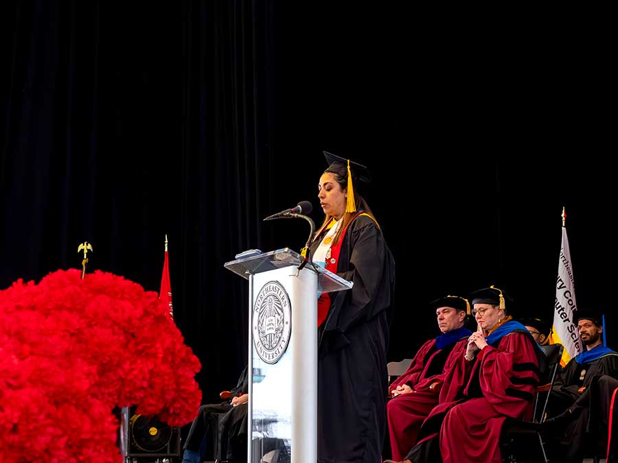 Khoury master’s student speaker Daniela Alejandra González speaks behind a podium with a large display of red flowers visible in front of the podium