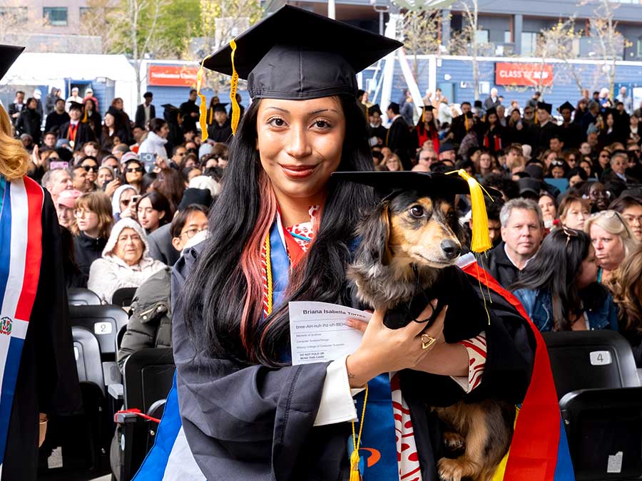 A Khoury graduate wearing a cap and gown holds her dog, which is also wearing a cap and gown