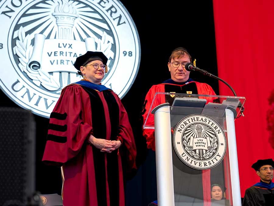 Elizabeth Mynatt (left) and Ben Hescott stand behind a podium while Hescott speaks with the Northeastern University seal hanging in the background
