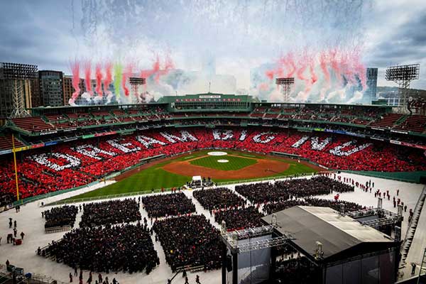 White letters that spell the phrase "Dream big NU" are visible in the lower level of Fenway Park during Northeastern's graduation ceremony as colored smoke is launched from the stadium's roof
