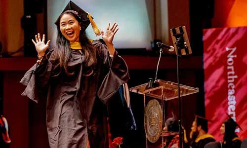 A Northeastern University Seattle graduate walks across a stage while raising her hands in celebration. Photo by Diwas Photography for Northeastern University