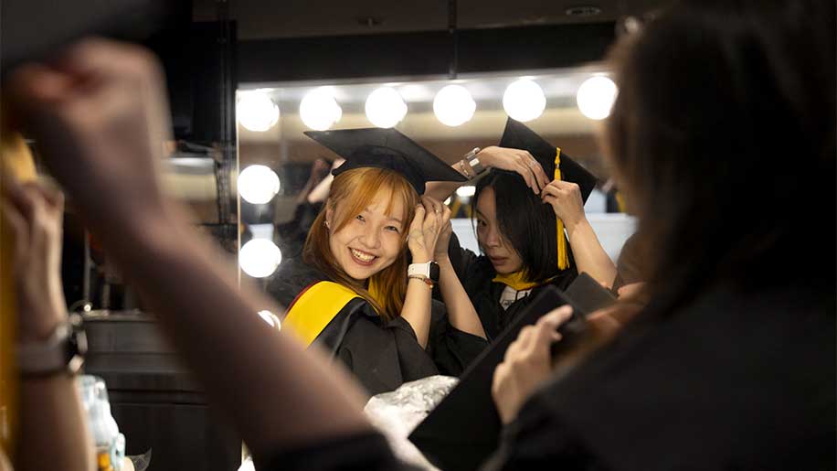 Two students adjust their graduation regalia while looking in a mirror