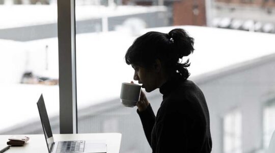 The silhouette of a person in front of a window sipping from a mug while sitting at a desk looking at a laptop