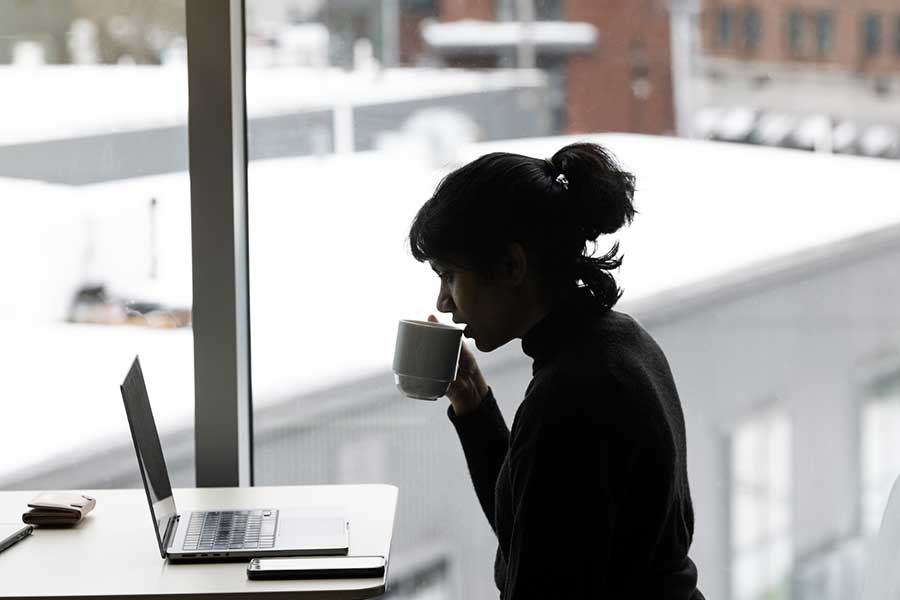 The silhouette of a person in front of a window sipping from a mug while sitting at a desk looking at a laptop