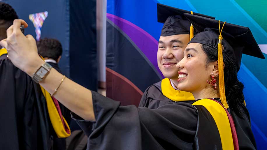 A Roux graduate wearing regalia takes a selfie with a fellow graduate.