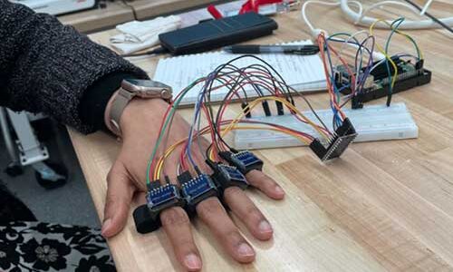 A hand is resting on a desk while four fingers are connected to wires