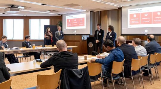 A student team presents their project on two large projection screens as an audience sitting at a large, u-shaped table watches