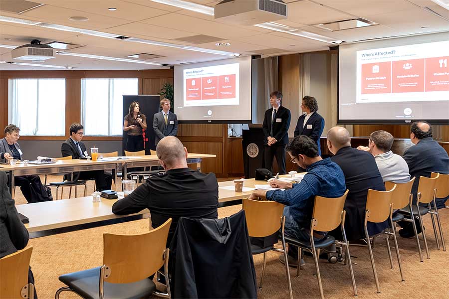 A student team presents their project on two large projection screens as an audience sitting at a large, u-shaped table watches