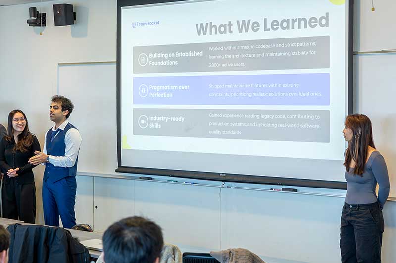 Three students discuss their project in front of a large presentation screen showing a slide titled, "What We Learned"