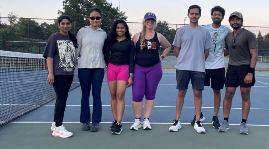 Seven members of the Roux tennis club pose for a photo on a tennis court next to the court's net