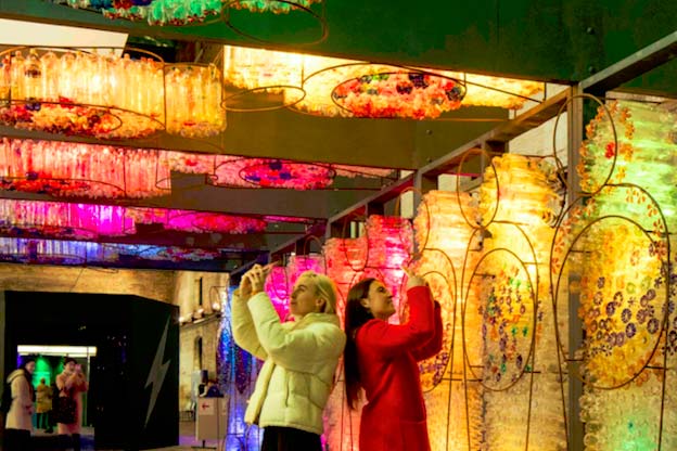 Two students stand back to back and take photos of a light and glass art exhibit