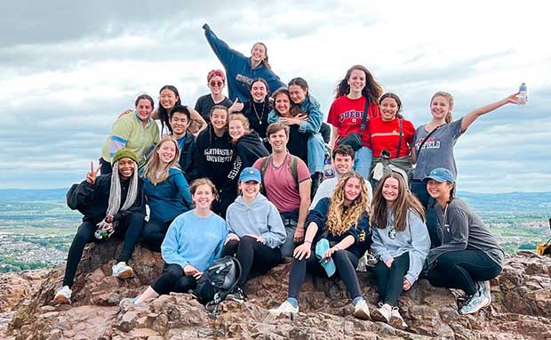 About 20 Northeastern students pose for a photo sitting and standing on top of a mountain in England.
